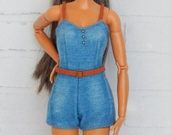 Può includere: Una bambola con i capelli castani che indossa una tuta di jeans blu con una cintura marrone. La bambola ha gli occhi marroni e indossa il trucco.