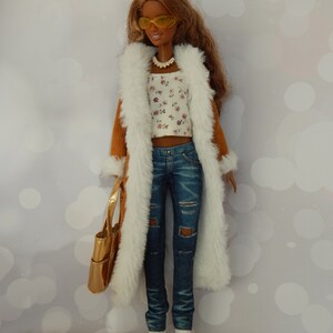 May include: A brown corduroy coat with a white faux fur lining, worn over a white floral top and blue jeans. The doll is wearing yellow sunglasses and a gold necklace.