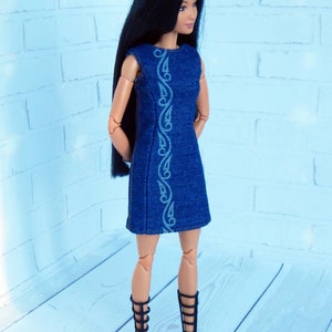May include: A doll wearing a blue denim dress with white embroidery and black gladiator sandals. The doll has long black hair and is standing on a light wood surface.