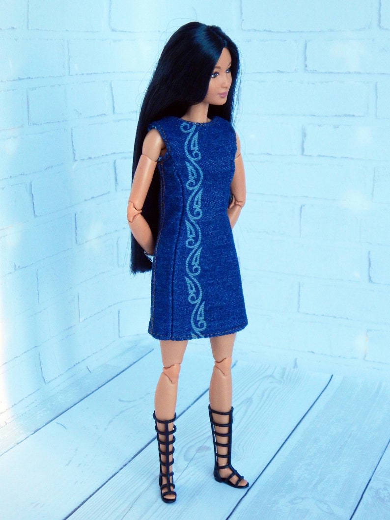 May include: A doll wearing a blue denim dress with white embroidery and black gladiator sandals. The doll has long black hair and is standing on a light wood surface.