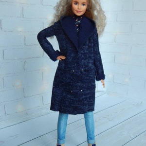 Pode incluir: Uma boneca vestindo um casaco azul de malha com uma gola grande, jeans azul e botas azuis. A boneca tem cabelo longo e loiro e olhos azuis.