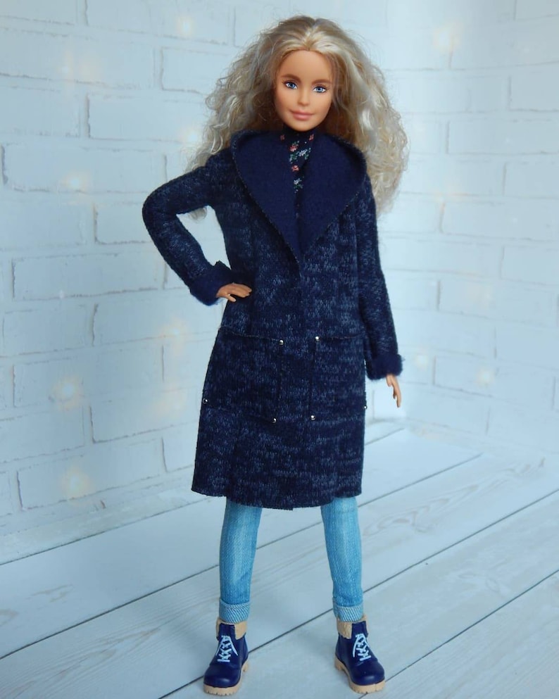 Pode incluir: Uma boneca vestindo um casaco azul de malha com uma gola grande, jeans azul e botas azuis. A boneca tem cabelo longo e loiro e olhos azuis.