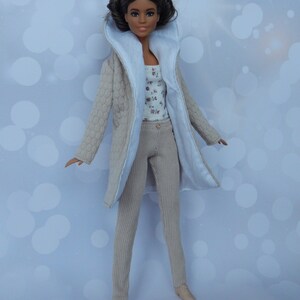 May include: A doll wearing a beige quilted coat with a white collar, beige pants, and a floral print top. The doll has brown hair and brown eyes.