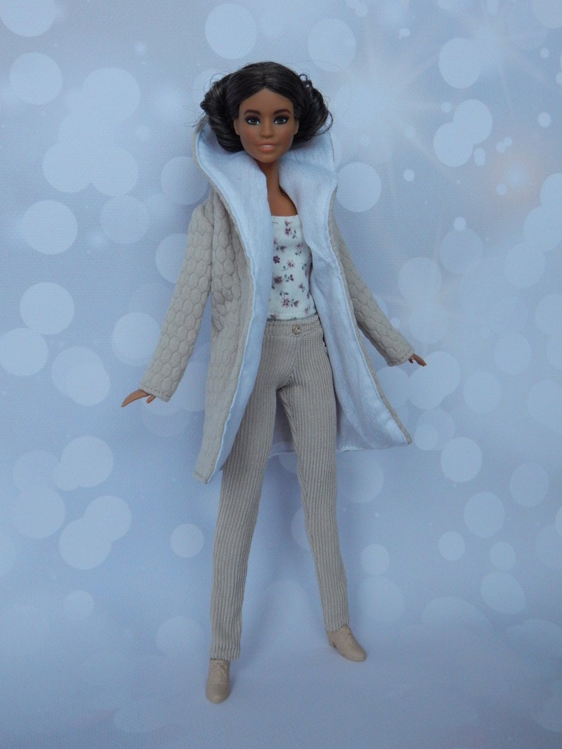 May include: A doll wearing a beige quilted coat with a white collar, beige pants, and a floral print top. The doll has brown hair and brown eyes.