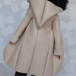 May include: A cream-colored faux leather coat with a large hood and a pleated skirt. The coat has a simple design and is made of a soft, supple material.