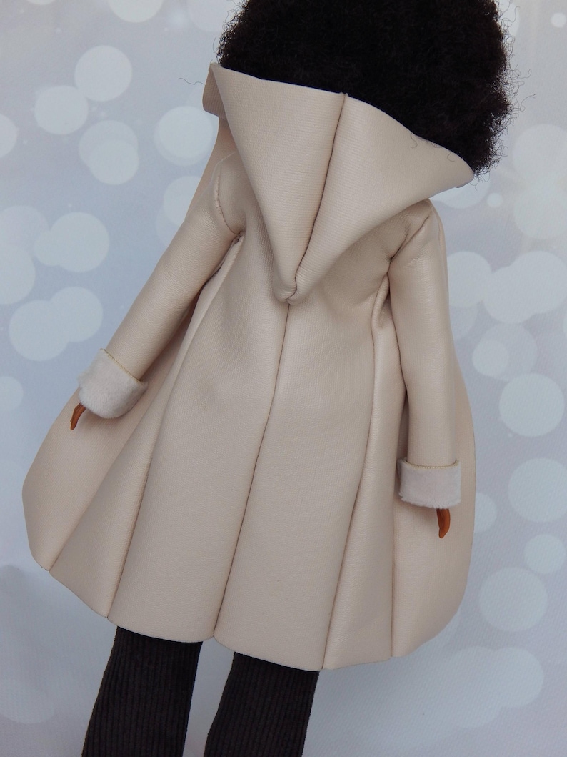 May include: A cream-colored faux leather coat with a large hood and a pleated skirt. The coat has a simple design and is made of a soft, supple material.