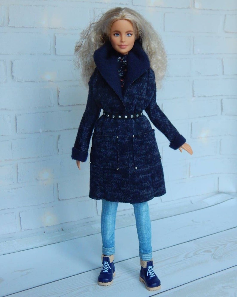 Pode incluir: Uma boneca vestindo um casaco azul com um cinto preto, jeans azul e botas azuis. A boneca tem cabelo loiro encaracolado e olhos azuis.