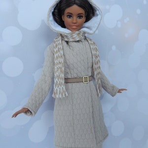 Puede incluir: Una mu&ntilde;eca con un abrigo beige con capucha, forro blanco, cintur&oacute;n marr&oacute;n y una bufanda blanca y marr&oacute;n. La mu&ntilde;eca tiene el pelo casta&ntilde;o y los ojos marrones.