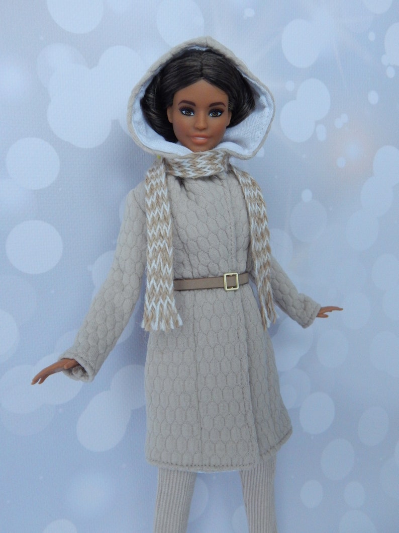 Puede incluir: Una mu&ntilde;eca con un abrigo beige con capucha, forro blanco, cintur&oacute;n marr&oacute;n y una bufanda blanca y marr&oacute;n. La mu&ntilde;eca tiene el pelo casta&ntilde;o y los ojos marrones.