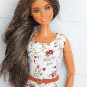 K&ouml;nnte beinhalten: Eine Puppe mit langen braunen Haaren, die ein wei&szlig;es Kleid mit Blumenmuster und einen braunen G&uuml;rtel tr&auml;gt.