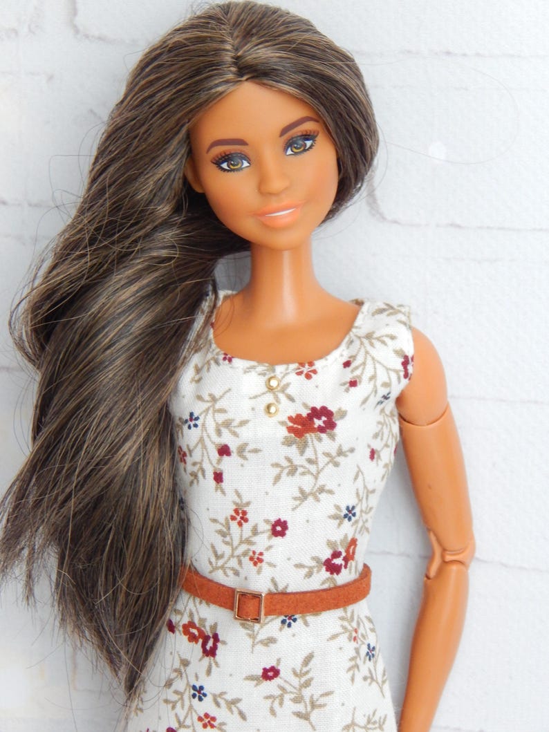 K&ouml;nnte beinhalten: Eine Puppe mit langen braunen Haaren, die ein wei&szlig;es Kleid mit Blumenmuster und einen braunen G&uuml;rtel tr&auml;gt.