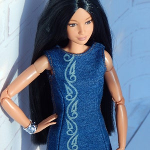 May include: A doll with long black hair wearing a blue denim dress with a white design. The doll has brown eyes and is standing against a white brick wall.