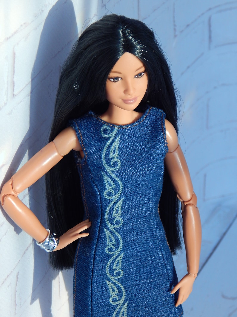 May include: A doll with long black hair wearing a blue denim dress with a white design. The doll has brown eyes and is standing against a white brick wall.