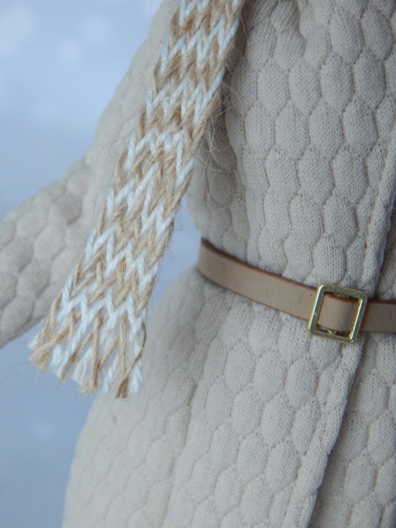 Puede incluir: Un su&eacute;ter de punto beige con un cintur&oacute;n de cuero marr&oacute;n y una bufanda tejida blanca y beige.