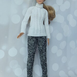 May include: A doll wearing a white turtleneck sweater and black and white patterned pants. The doll has long blonde hair and blue eyes.