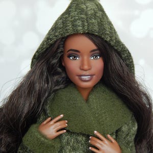 May include: A doll with long dark hair wearing a green knit hooded coat with a matching collar and a pom-pom on the hood. The doll has brown eyes and is smiling. The coat has a textured appearance.