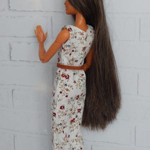 K&ouml;nnte beinhalten: Eine Puppe tr&auml;gt ein wei&szlig;es Kleid mit Blumenmuster. Das Kleid hat einen braunen G&uuml;rtel und die Puppe hat lange braune Haare.