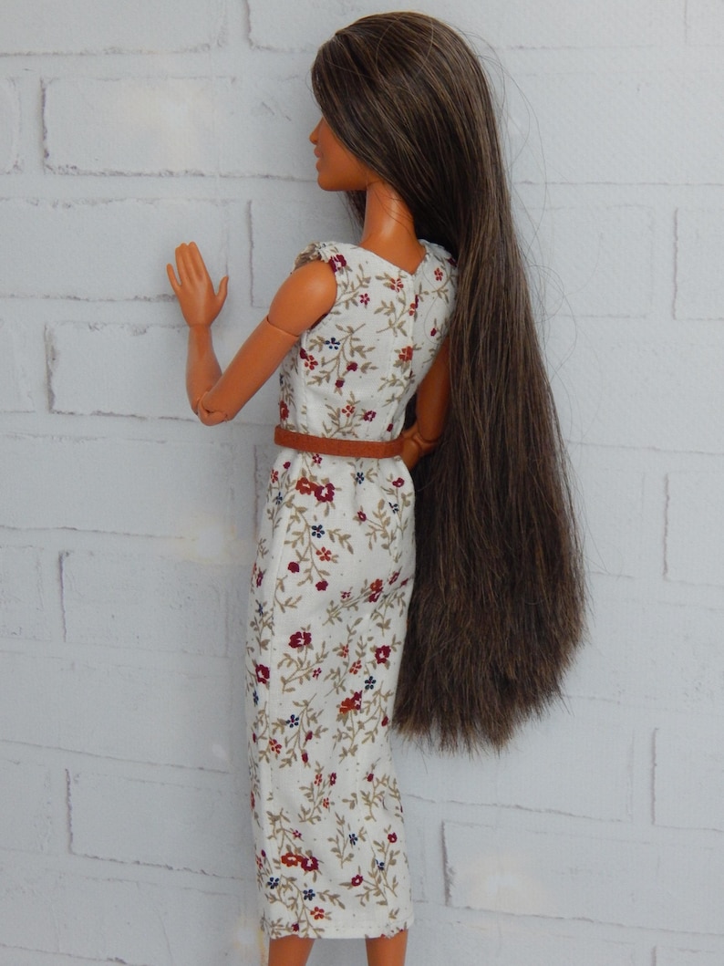K&ouml;nnte beinhalten: Eine Puppe tr&auml;gt ein wei&szlig;es Kleid mit Blumenmuster. Das Kleid hat einen braunen G&uuml;rtel und die Puppe hat lange braune Haare.