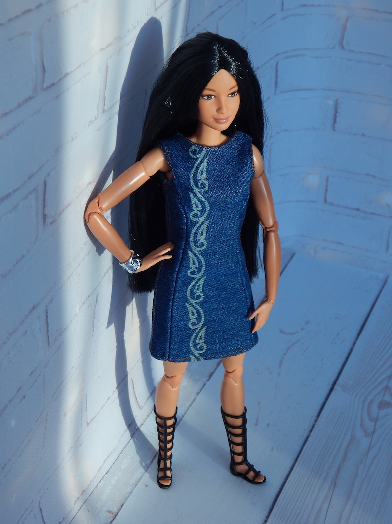 May include: A fashion doll with long black hair wearing a blue denim dress with a white design and black gladiator sandals.