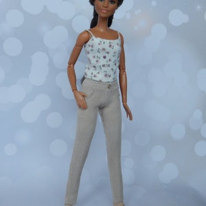 May include: A doll with long dark brown hair wearing a white tank top with a floral print and beige corduroy pants.
