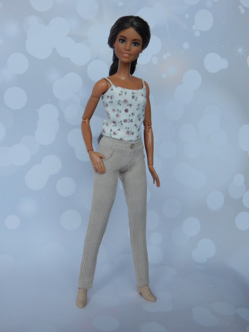May include: A doll with long dark brown hair wearing a white tank top with a floral print and beige corduroy pants.