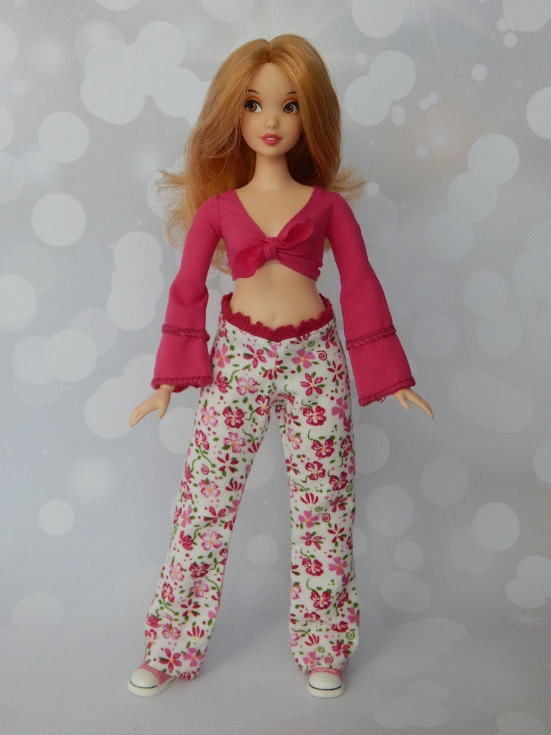 May include: A doll wearing a pink crop top with a bow and white pants with a floral pattern.