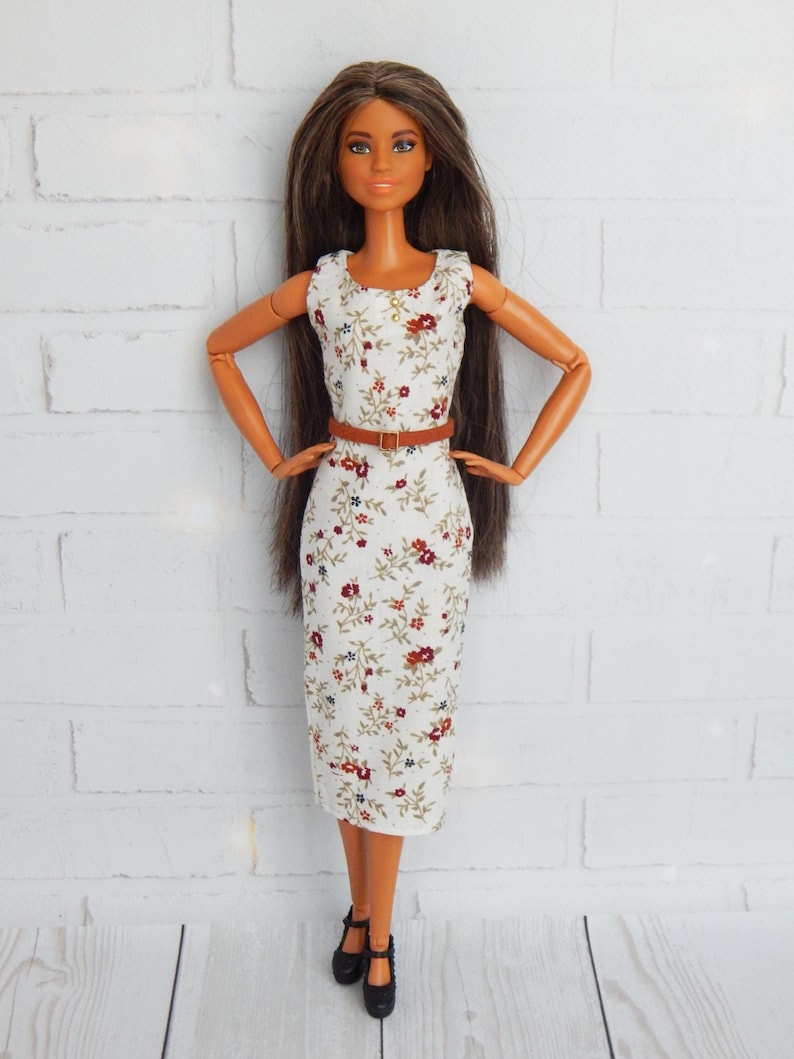 K&ouml;nnte beinhalten: Eine Puppe, die ein wei&szlig;es Kleid mit Blumenmuster und einen braunen G&uuml;rtel tr&auml;gt. Die Puppe hat lange braune Haare und braune Augen.