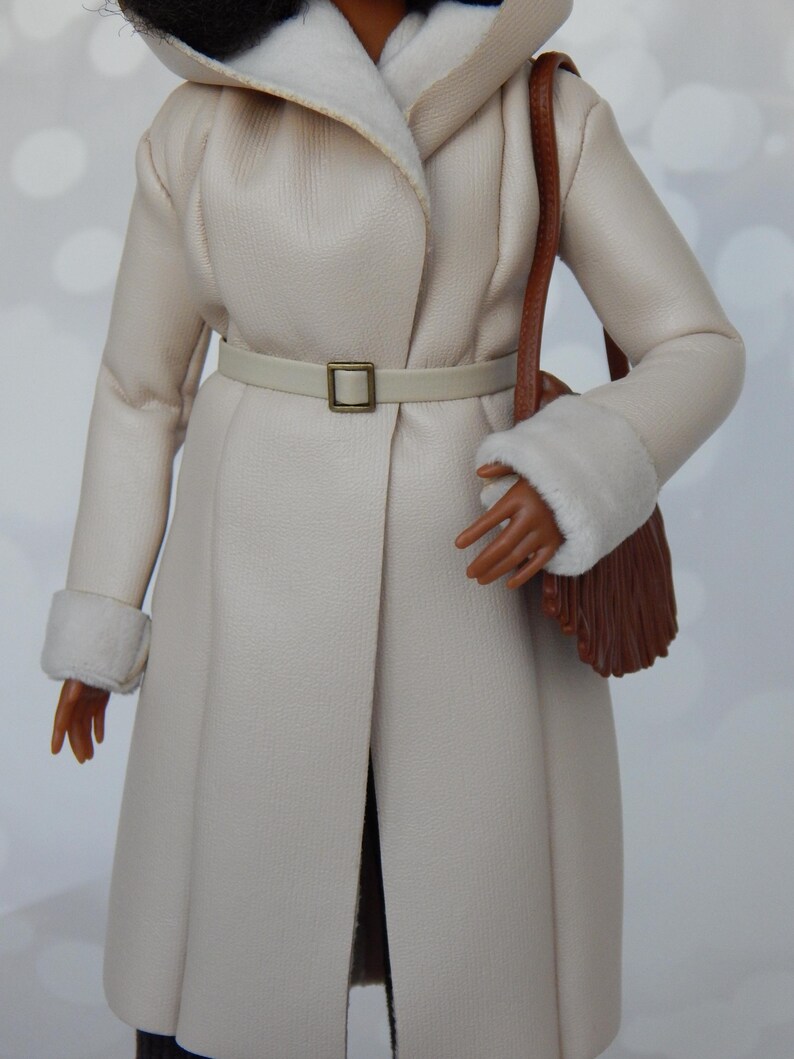 May include: A white faux fur coat with a brown leather strap and a brown fringe bag. The coat has a white fur collar and cuffs. The coat is belted with a white leather belt with a gold buckle.