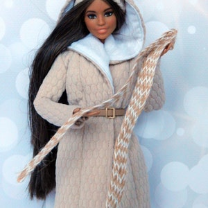 Puede incluir: Una mu&ntilde;eca con un abrigo beige con capucha, ribete de piel blanca, una bufanda marr&oacute;n y blanca y un cintur&oacute;n marr&oacute;n. La mu&ntilde;eca tiene el pelo largo y negro y los ojos marrones.