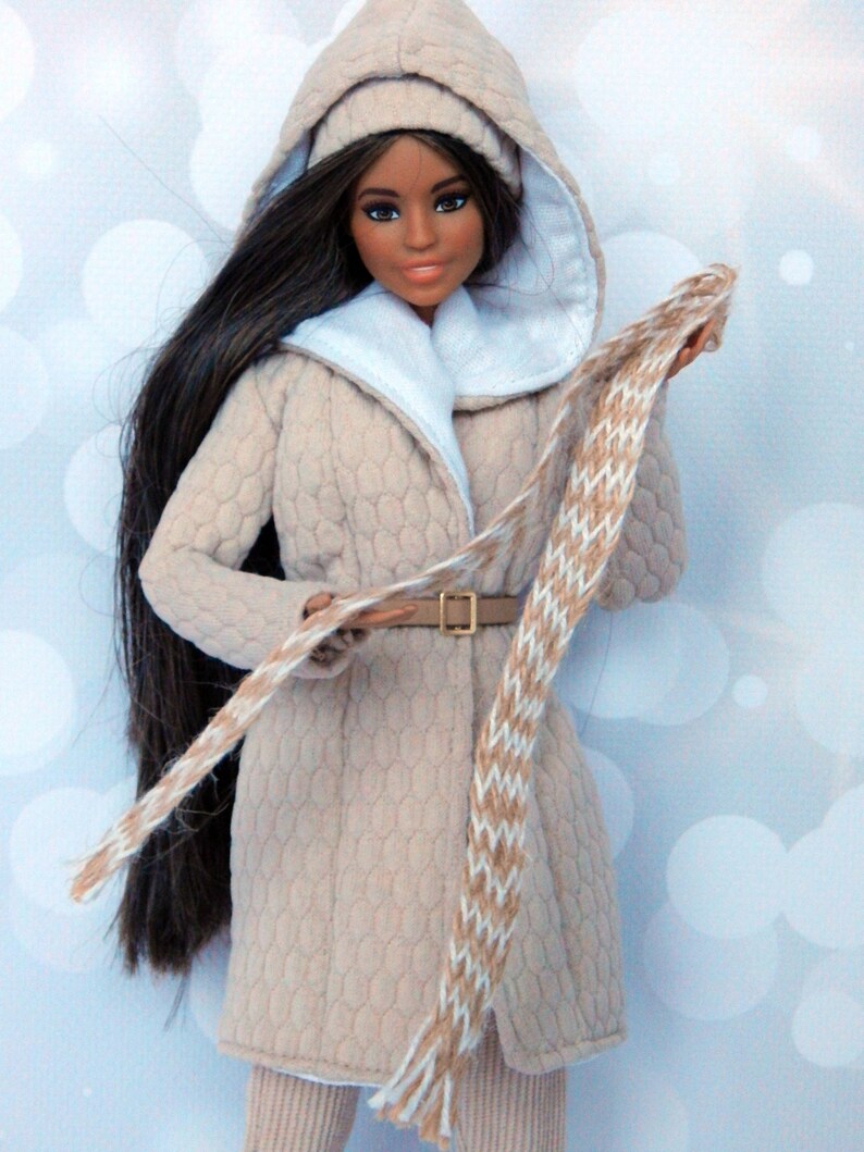 Puede incluir: Una mu&ntilde;eca con un abrigo beige con capucha, ribete de piel blanca, una bufanda marr&oacute;n y blanca y un cintur&oacute;n marr&oacute;n. La mu&ntilde;eca tiene el pelo largo y negro y los ojos marrones.