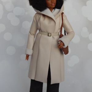 May include: A doll wearing a cream-colored faux fur coat with a brown leather belt and a brown leather bag. The doll has black hair styled in an afro and is wearing black pants.