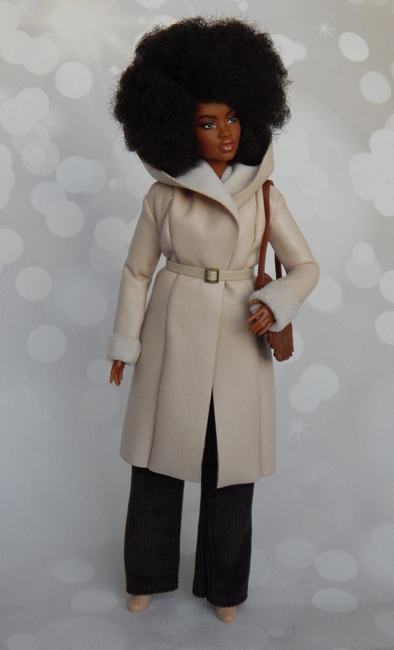 May include: A doll wearing a cream-colored faux fur coat with a brown leather belt and a brown leather bag. The doll has black hair styled in an afro and is wearing black pants.