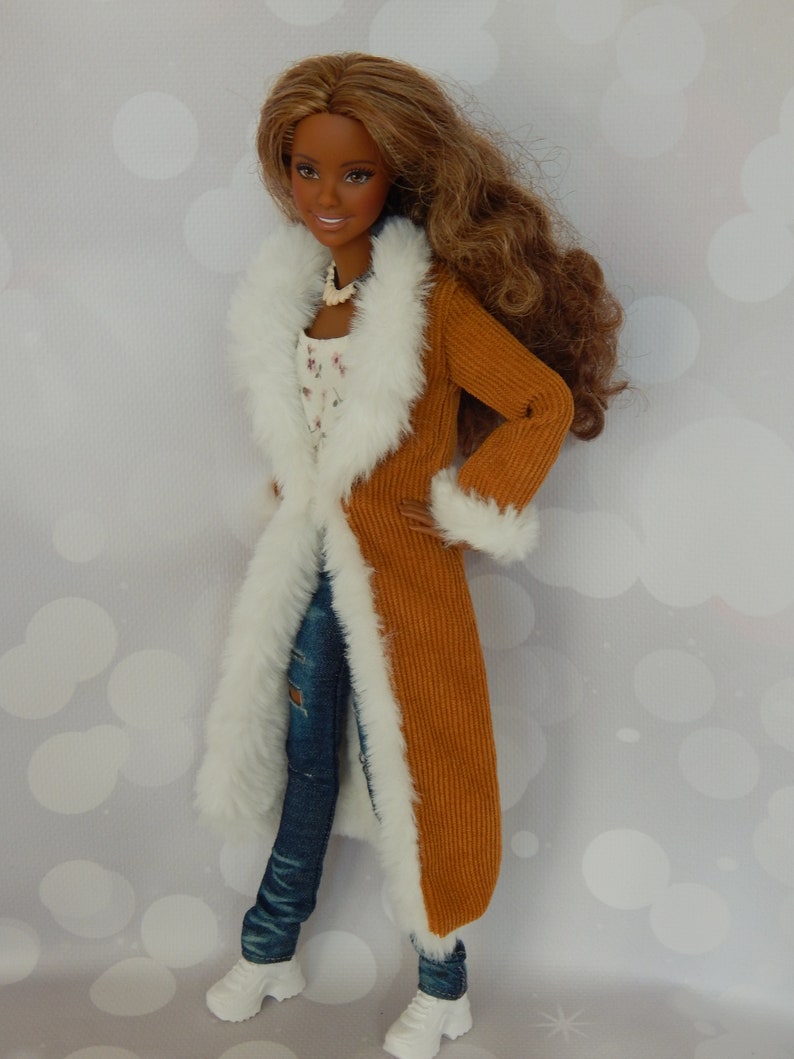 May include: A doll wearing a brown corduroy coat with a white fur trim, blue jeans, and white sneakers. The doll has brown hair and brown eyes.