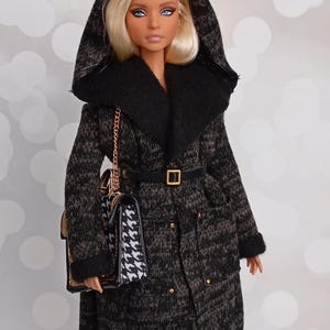 May include: A doll wearing a black and gray patterned coat with a hood and a black belt. The doll has blonde hair and is carrying a black and white handbag with a gold chain. The coat has gold buttons and a black collar.