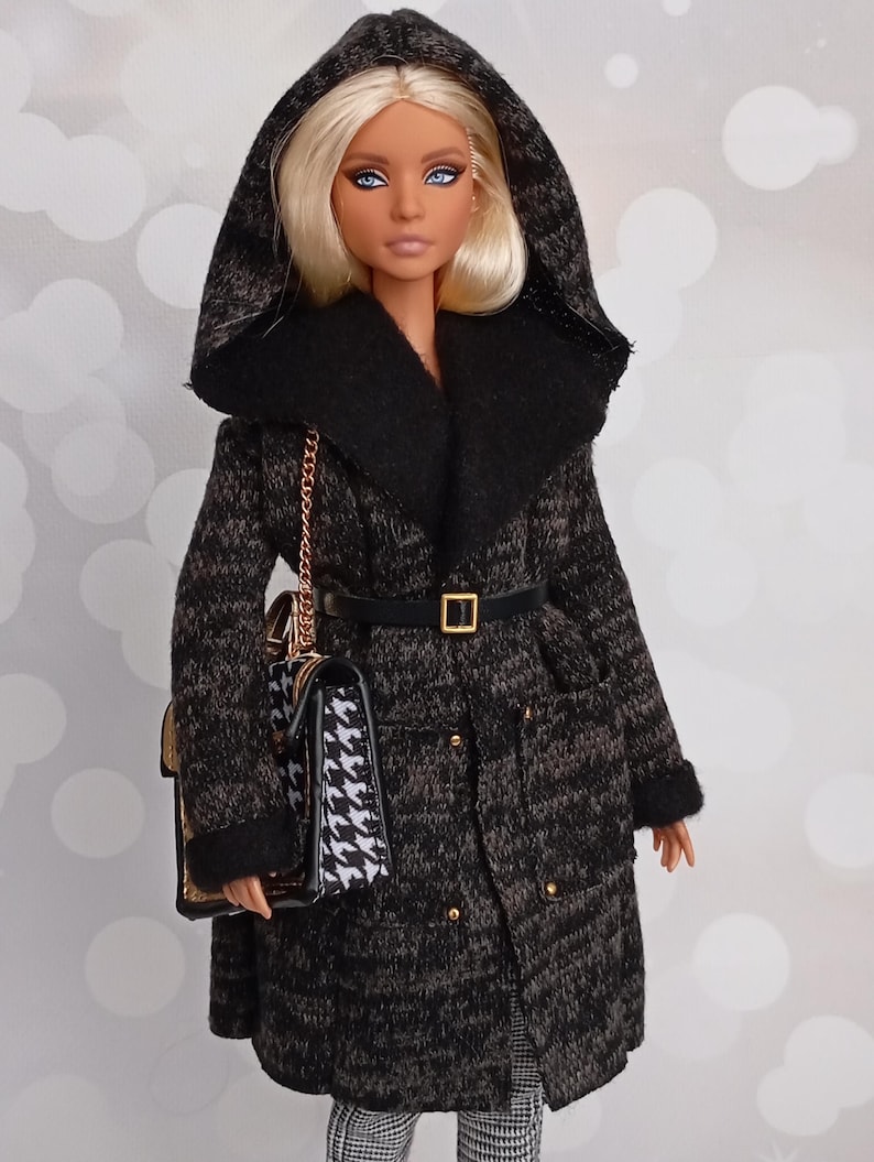 May include: A doll wearing a black and gray patterned coat with a hood and a black belt. The doll has blonde hair and is carrying a black and white handbag with a gold chain. The coat has gold buttons and a black collar.