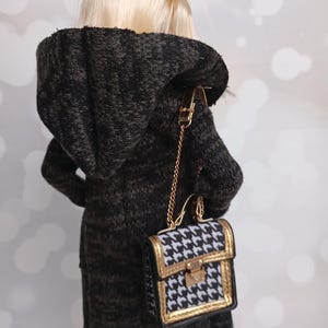 May include: A doll wearing a black and gray hooded coat and carrying a small purse. The purse has a black and white houndstooth pattern with gold trim and a gold chain strap. The doll has blonde hair.
