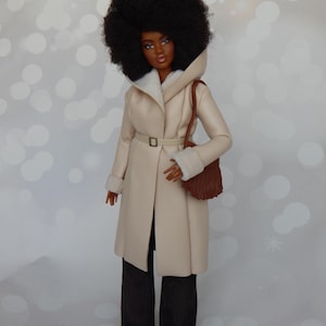 May include: A doll wearing a cream-colored coat with a brown belt and a brown purse. The doll has brown eyes, dark brown hair, and is wearing brown pants.