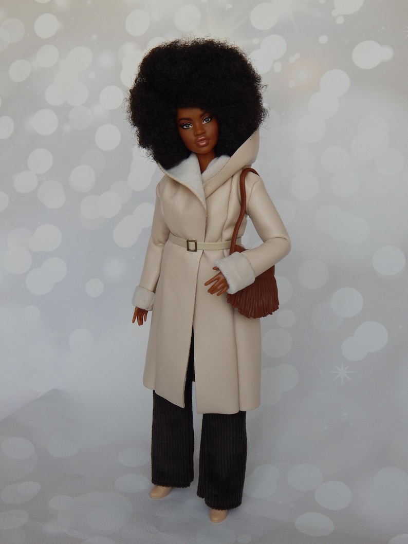 May include: A doll wearing a cream-colored coat with a brown belt and a brown purse. The doll has brown eyes, dark brown hair, and is wearing brown pants.