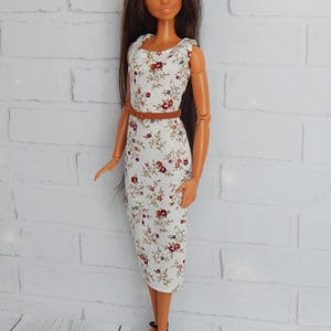 K&ouml;nnte beinhalten: Eine Puppe, die ein wei&szlig;es Kleid mit Blumenmuster und einen braunen G&uuml;rtel tr&auml;gt. Die Puppe hat lange braune Haare und braune Augen. Die Puppe tr&auml;gt schwarze Schuhe.