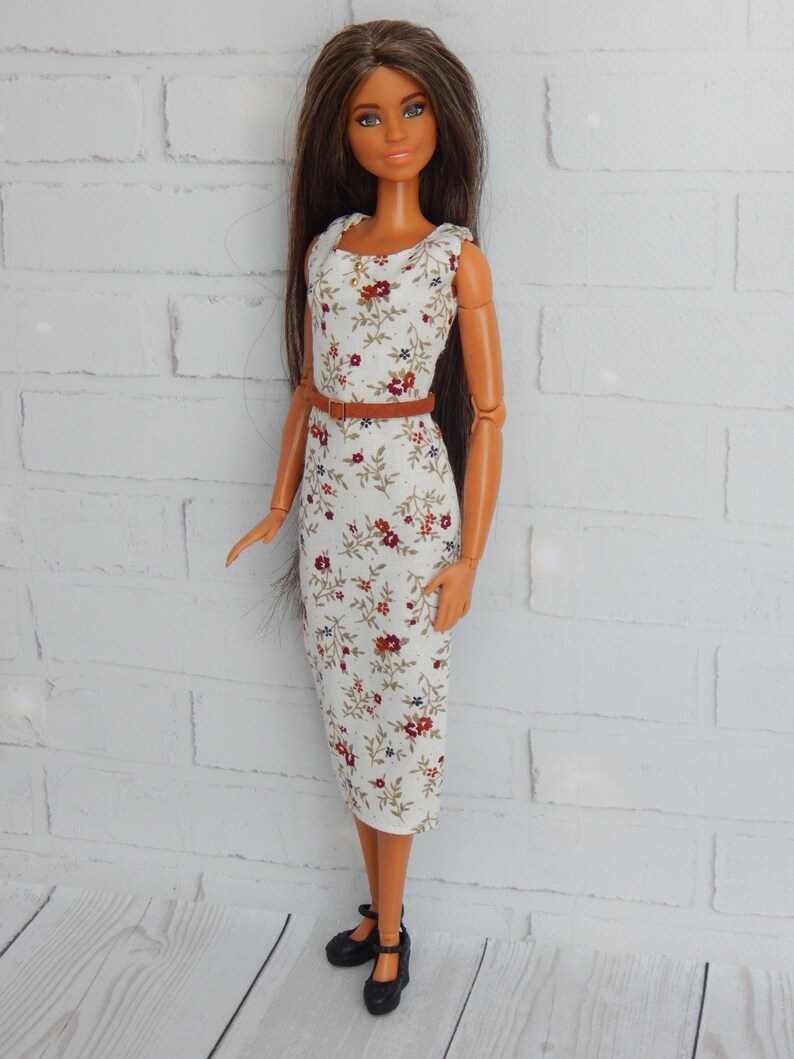 K&ouml;nnte beinhalten: Eine Puppe, die ein wei&szlig;es Kleid mit Blumenmuster und einen braunen G&uuml;rtel tr&auml;gt. Die Puppe hat lange braune Haare und braune Augen. Die Puppe tr&auml;gt schwarze Schuhe.