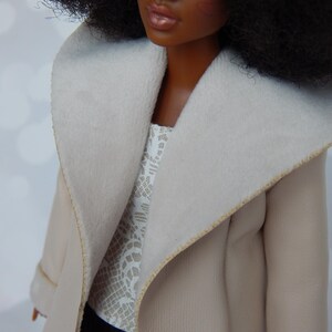 May include: A doll wearing a beige coat with a white fur collar and a white lace top. The doll has dark brown skin, black hair, and brown eyes.