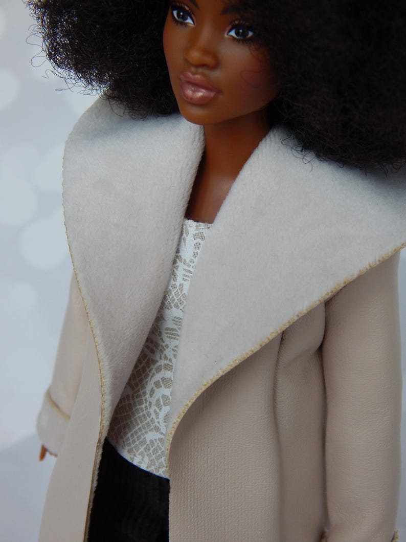 May include: A doll wearing a beige coat with a white fur collar and a white lace top. The doll has dark brown skin, black hair, and brown eyes.