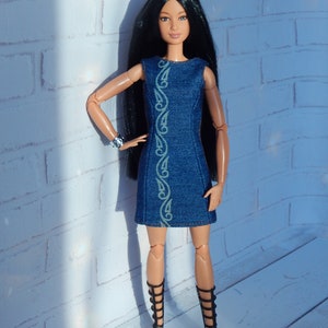 May include: A black-haired doll wearing a blue denim dress with a green floral design and black gladiator sandals.