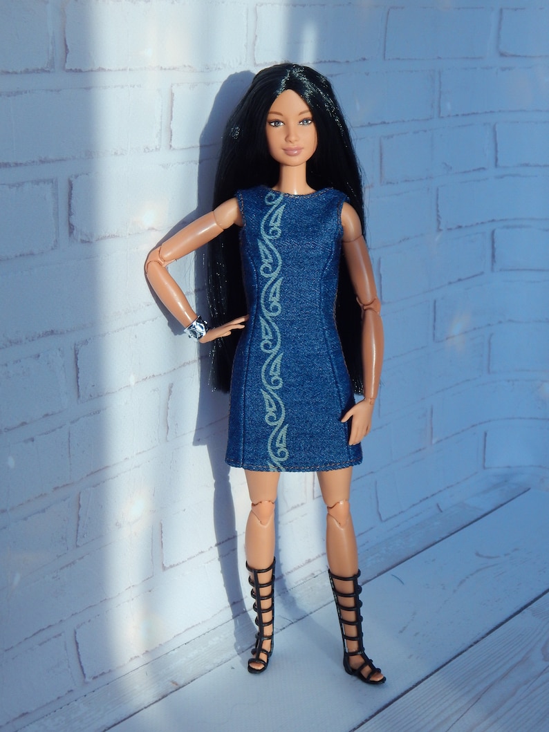 May include: A black-haired doll wearing a blue denim dress with a green floral design and black gladiator sandals.