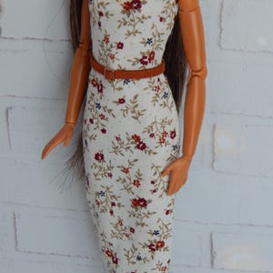 K&ouml;nnte beinhalten: Ein wei&szlig;es, &auml;rmelloses Kleid mit einem Blumendruck in Rot-, Braun- und Blaut&ouml;nen. Das Kleid hat einen braunen G&uuml;rtel und ist figurbetont.