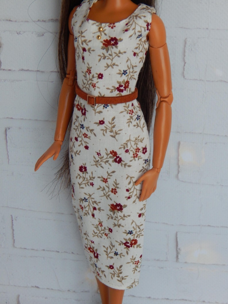 K&ouml;nnte beinhalten: Ein wei&szlig;es, &auml;rmelloses Kleid mit einem Blumendruck in Rot-, Braun- und Blaut&ouml;nen. Das Kleid hat einen braunen G&uuml;rtel und ist figurbetont.