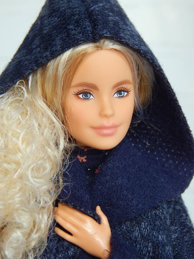 Pode incluir: Uma boneca loira com olhos azuis vestindo um su&eacute;ter azul escuro com capuz.