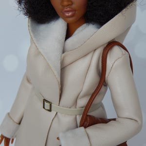 May include: A doll wearing a cream-colored faux fur hooded coat with a brown leather belt and a brown fringed bag.