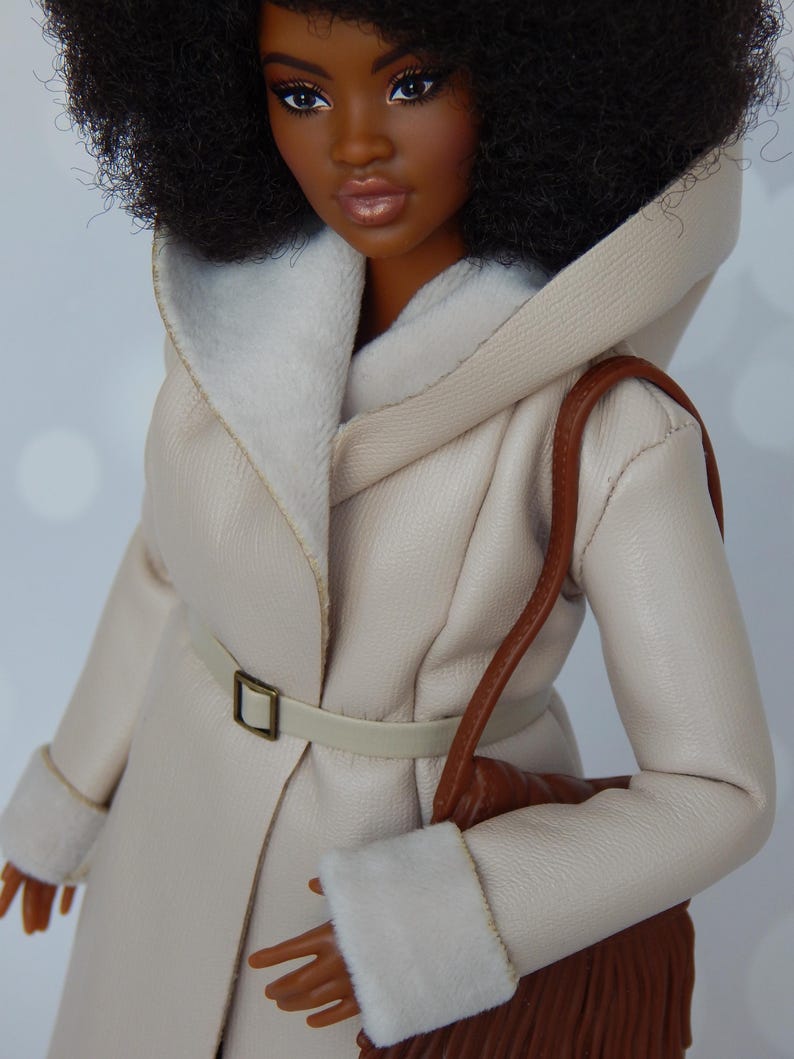 May include: A doll wearing a cream-colored faux fur hooded coat with a brown leather belt and a brown fringed bag.