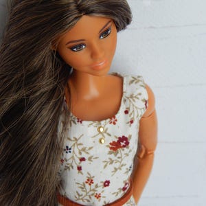 K&ouml;nnte beinhalten: Eine Puppe mit langen braunen Haaren, die ein wei&szlig;es Kleid mit Blumenmuster und einen braunen G&uuml;rtel tr&auml;gt.